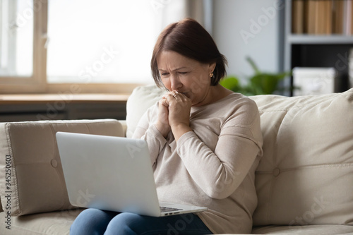 Sad mature woman looking at laptop screen and crying, sitting on couch at home, unhappy older female using computer, reading bad news, debt or bankruptcy, watching movie, making video call