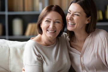 Happy mature mother and adult daughter hugging close up, having fun, sitting on couch at home, smiling woman embracing older mum, spending leisure time together, two generations good relationship