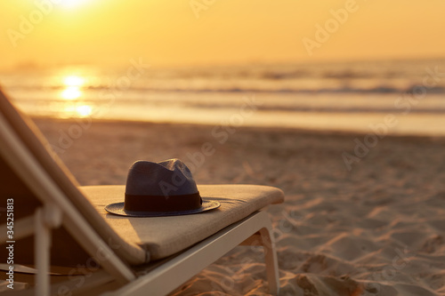 Fotografija Blue hat is lying on a chaise longue on a beautiful sandy beach by the sea in the rays of the setting sun
