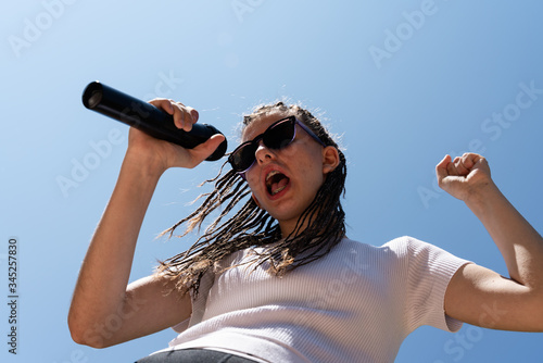 White teenager with hair braids wearing a white top and sun glasses singing a song with a black microphone at the right hand and the sky and the sunlight at the background. Horizontal photo