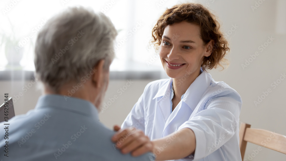Smiling female doctor reassuring supporting senior adult patient in hospital. Kind caring young woman nurse or caregiver helping older retired man talking, giving comfort, expressing care concept. Smiling female doctor reassuring supporting senior adult patient in hospital. Kind caring young woman nurse or caregiver helping older retired man talking, giving comfort, expressing care concept.