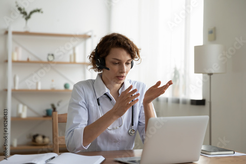 Foto Female general practitioner wears white coat and headset speaking videoconferencing on laptop computer using online video call consultation app