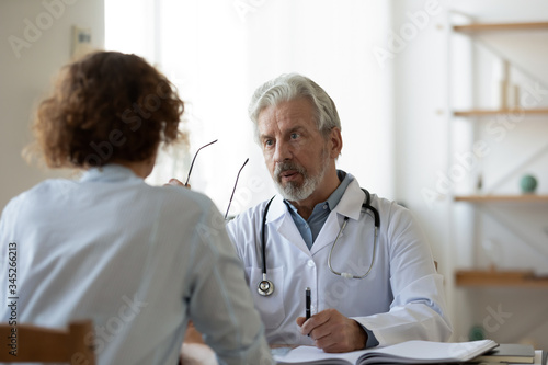 Professional senior male physician consulting female patient in hospital. Old doctor examining young adult woman at medical visit. Senior man therapist talking to client at healthcare checkup meeting