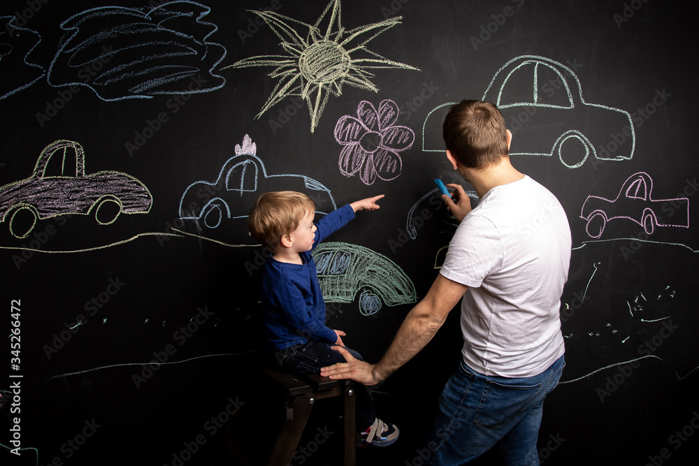 a little boy stands on the stairs near the slate wall and draws cars with his dad with chalk. children's creativity. home leisure with parents. studying at school with a teacher