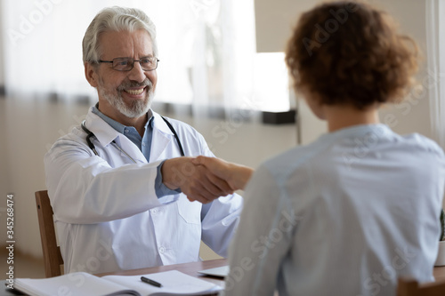 Friendly smiling professional physician handshaking female patient at medical consultation. Happy old male doctor greeting woman client shake hands express trust and gratitude for healthcare service.