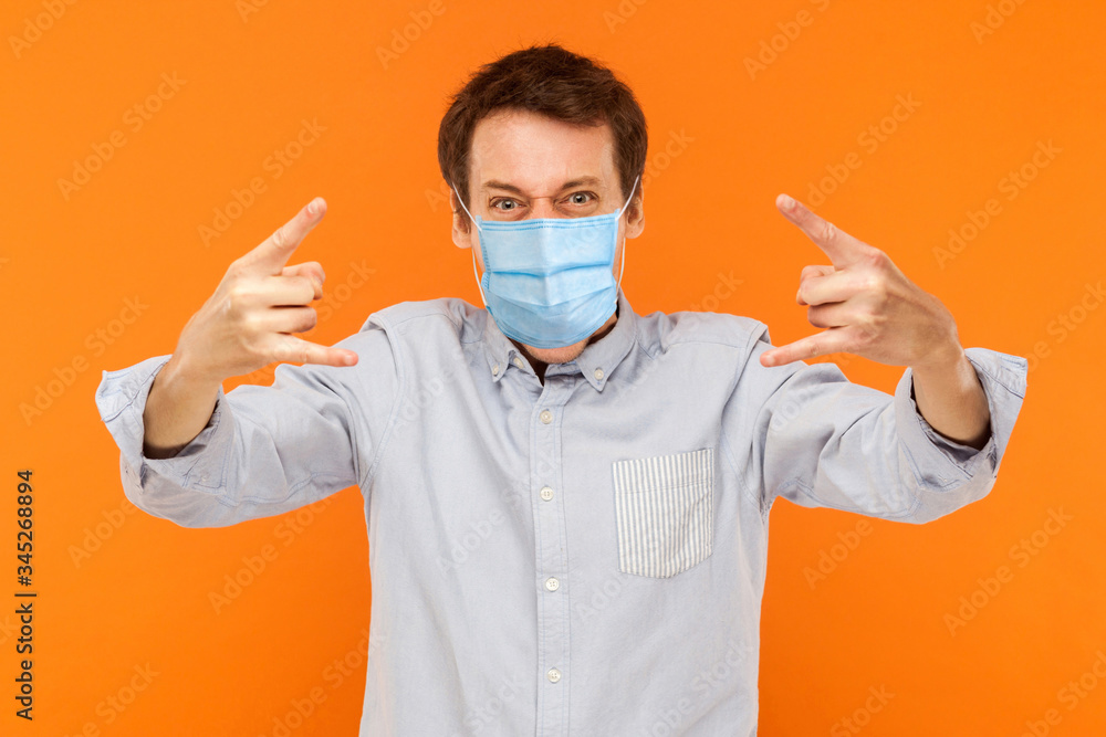 Yes. Portrait of young worker man with surgical medical mask standing with rock horns and loolong at camera screaming. indoor studio shot isolated on orange background. Yes. Portrait of young worker man with surgical medical mask standing with rock horns and loolong at camera screaming. indoor studio shot isolated on orange background.