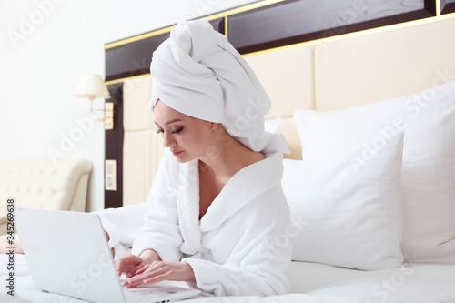 A young girl on the bed in a Bathrobe and a towel on her head is working on a laptop. The concept of distance learning or remote work. Home environment. Copy of the space.