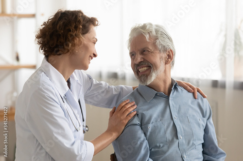 Cheerful healthy old male patient and female doctor having good trust relationship talking, bonding, laughing during medical visit. Happy physician helping, embracing, encouraging senior elder client.