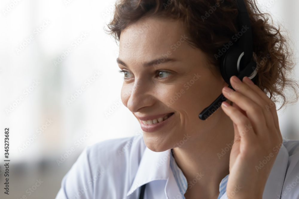 Close up head shot of smiling female young doctor operator wearing headset with microphone looking away. Medical call center healthcare services, telehealth hotline, remote tele medicine concept. Close up head shot of smiling female young doctor operator wearing headset with microphone looking away. Medical call center healthcare services, telehealth hotline, remote tele medicine concept.