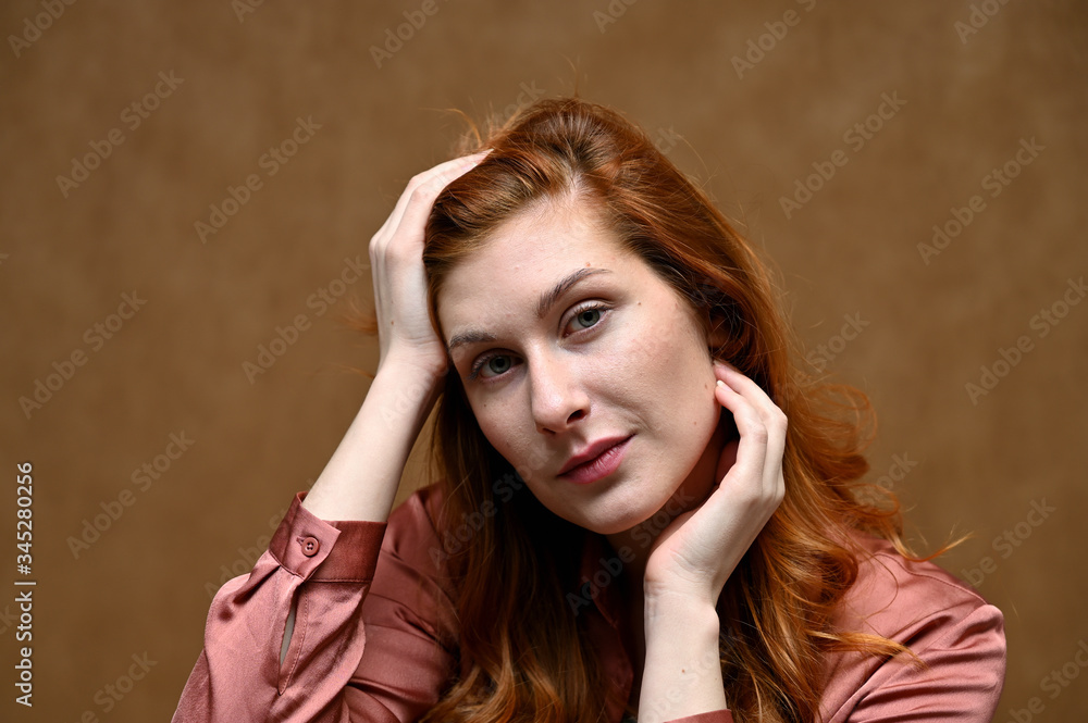 Obraz premium Close-up portrait of a young pretty woman with long red hair in a pink blouse. Photo taken on a beige background in the studio.