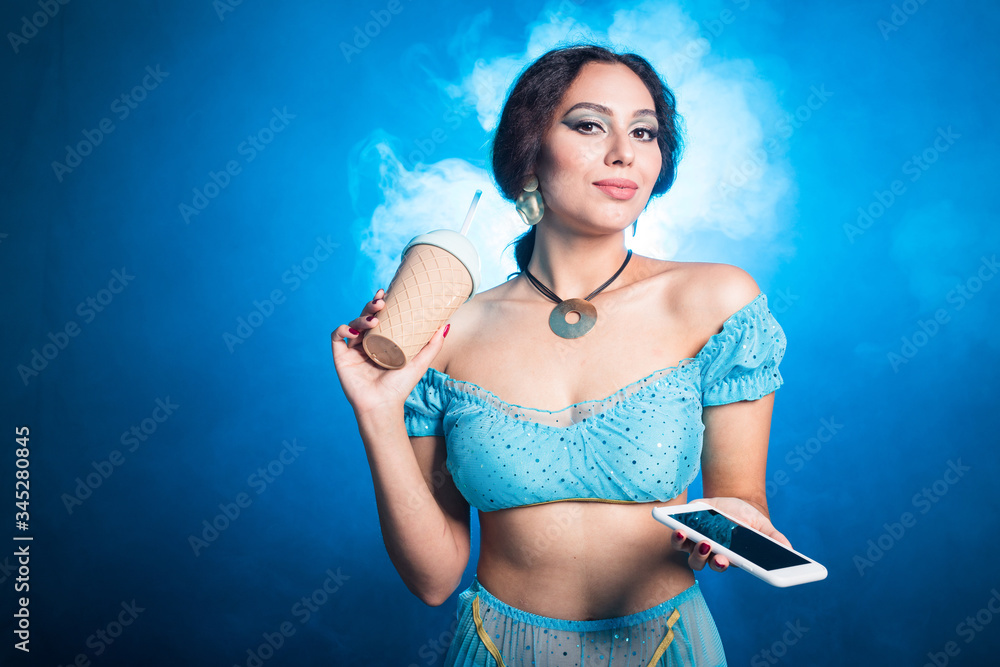 Obraz premium Portrait of young woman in the image of an Eastern fairy Princess in modern clothes holds smartphone and shaker. Carnival and halloween concept.