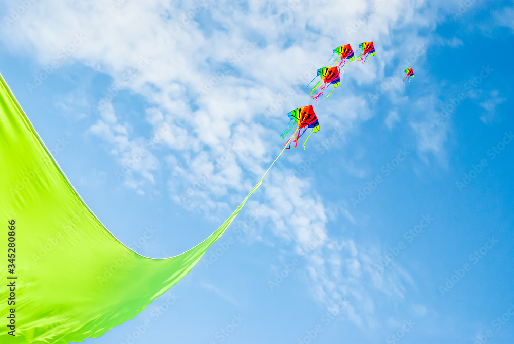 Rainbow colored kites flying high on the end of a vibrant green ribbon against a Summer blue sky and light clouds