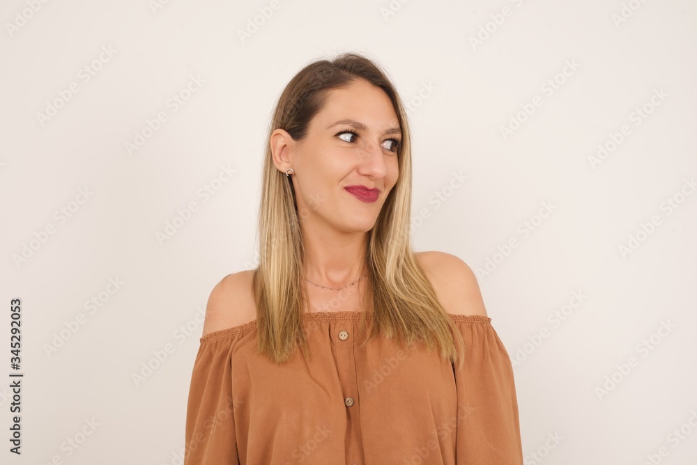 Dreamy young female with thoughtful expression, looks away keeps hands down bitting her lip thinks about something pleasant poses against gray wall. Dreamy young female with thoughtful expression, looks away keeps hands down bitting her lip thinks about something pleasant poses against gray wall.