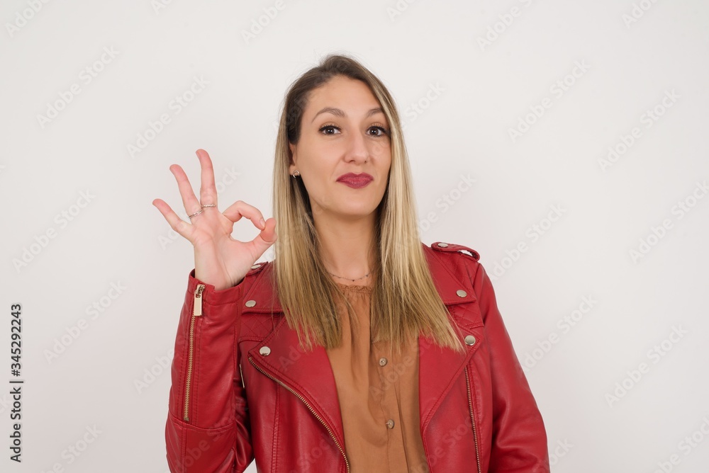 Glad attractive woman shows ok sign with hand as expresses approval, has cheerful expression, being optimistic. Standing against white wall. Glad attractive woman shows ok sign with hand as expresses approval, has cheerful expression, being optimistic. Standing against white wall.