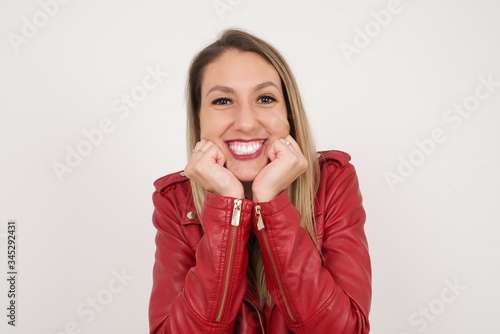 Dreamy young European woman keeps hands pressed together under chin, looks with happy expression, has toothy smile, isolated over gray background