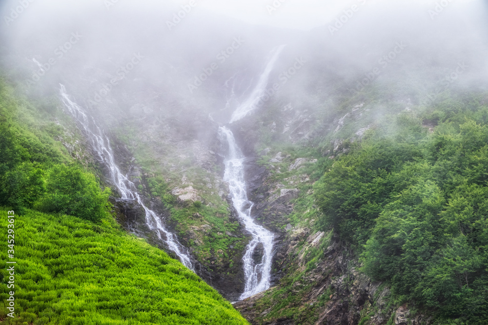Green mountain with a waterfall in spring or summer is covered in fog.
