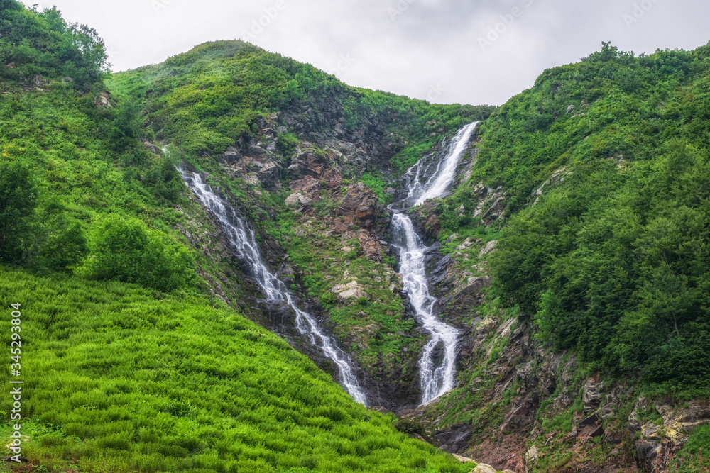 Green mountain range with a waterfall in spring or summer. A mountain range with a waterfall is covered in fog and clouds.