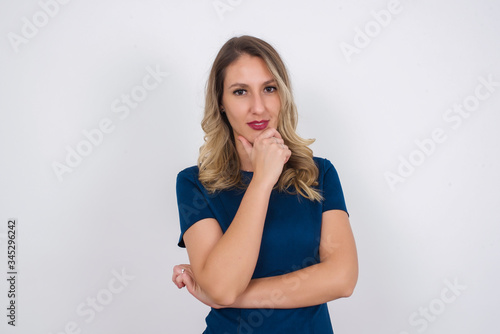 Portrait of thoughtful smiling girl keeps hand under chin, looks directly at camera, listens something with interest, dressed casually, poses against gray wall. Youth concept.