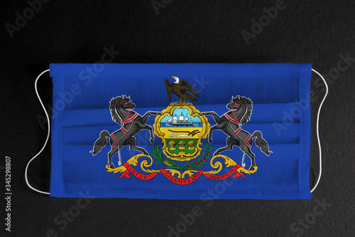 Coronavirus Covid-19 in Pennsylvania U.S. state. Flag of the State of Pennsylvania printed on medical mask on black background. Coronavirus update in Philadelphia. State healthcare concept.
 photo