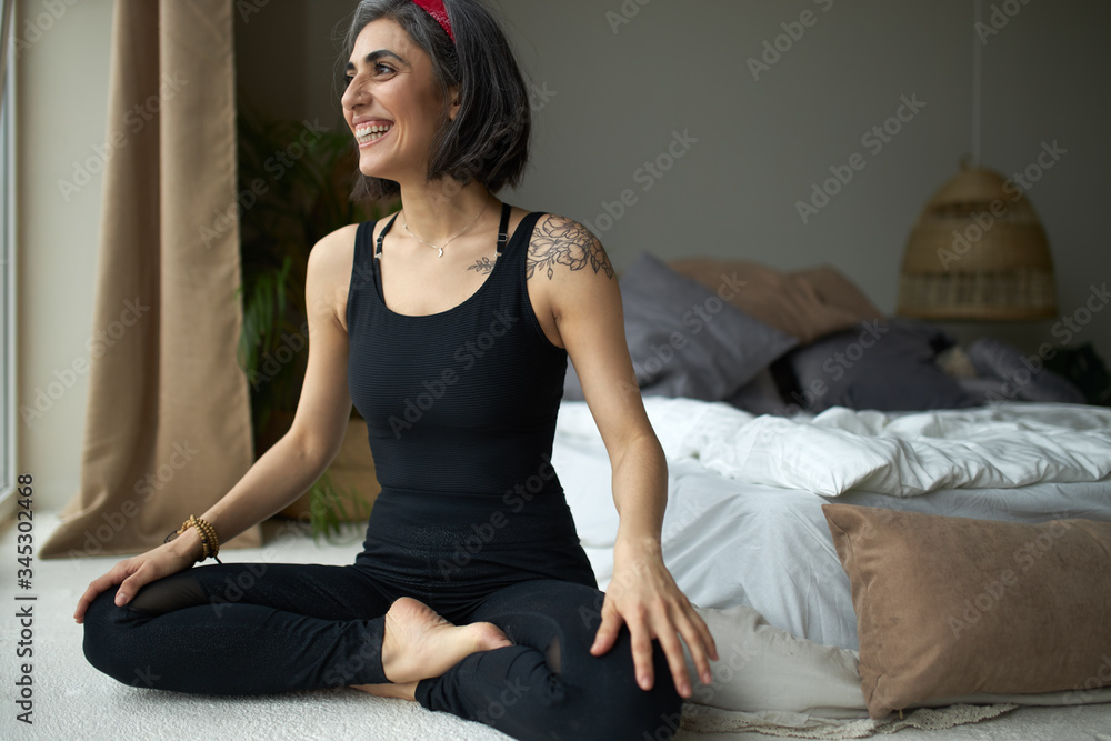 Stylish cute young female yoga instructor with slim fit body and grayish hair wearing black sports outfit doing advanced asanas sitting in padmasana or lotus pose, smiling broadly, having joyful look Stylish cute young female yoga instructor with slim fit body and grayish hair wearing black sports outfit doing advanced asanas sitting in padmasana or lotus pose, smiling broadly, having joyful look