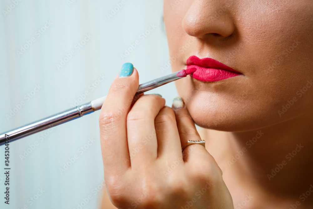 Makeup artist applies red lipstick. Beautiful female face. Hand of a make-up master painting lips of a young girl beauty model. Makeup in progress