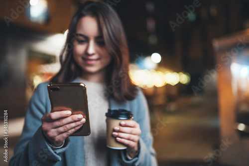 hipster girl drink coffee outdoors using mobile smartphone on background bokeh light in night city  woman hold in hands sellphone  online wi-fi internet