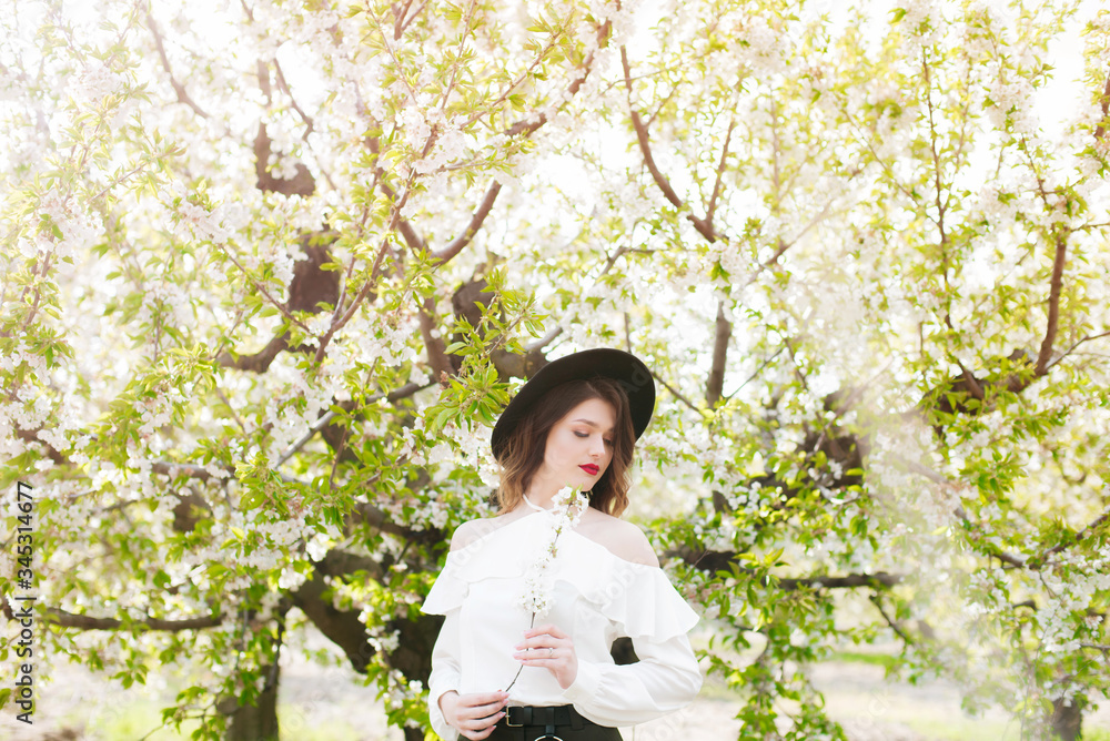Lovely young girl in a black hat and a white romantic silk blouse in spring in a blooming cherry garden in sunny weather with a blooming cherry branch in her hands. Spring and Fashion Lovely young girl in a black hat and a white romantic silk blouse in spring in a blooming cherry garden in sunny weather with a blooming cherry branch in her hands. Spring and Fashion