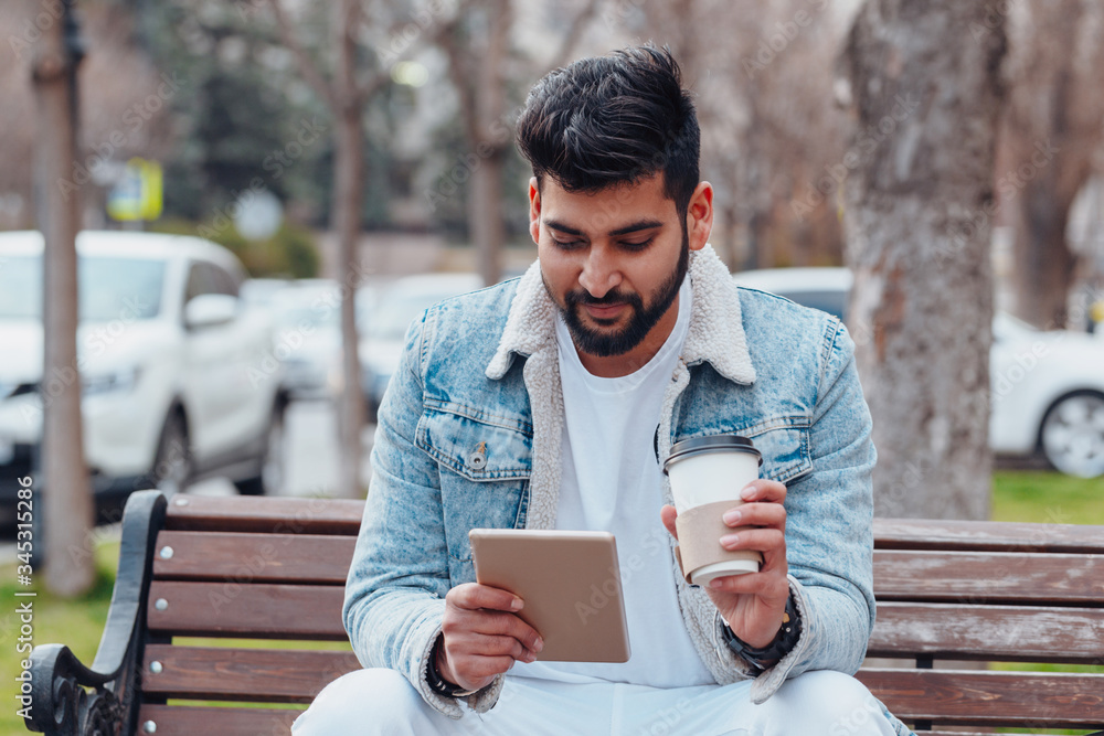Fototapeta premium Bearded Indian man sitting on a bench and using his tablet.