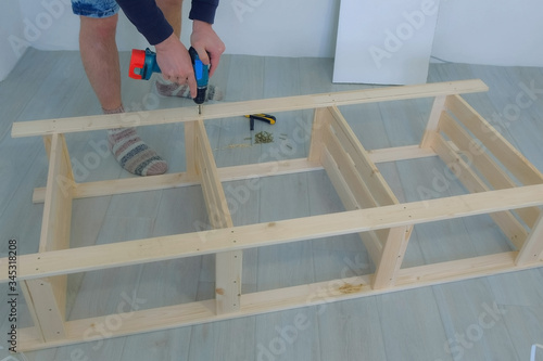 Man screwing boards with bolts using electric screwdriver collecting rack furniture bookcase, hands closeup. Assembling wooden rack on floor at home house. Moving at new place. Guy making renovation.