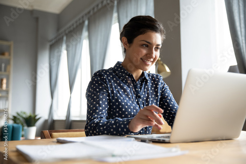 Photography Head shot smiling pretty indian girl sitting at table, looking at laptop screen