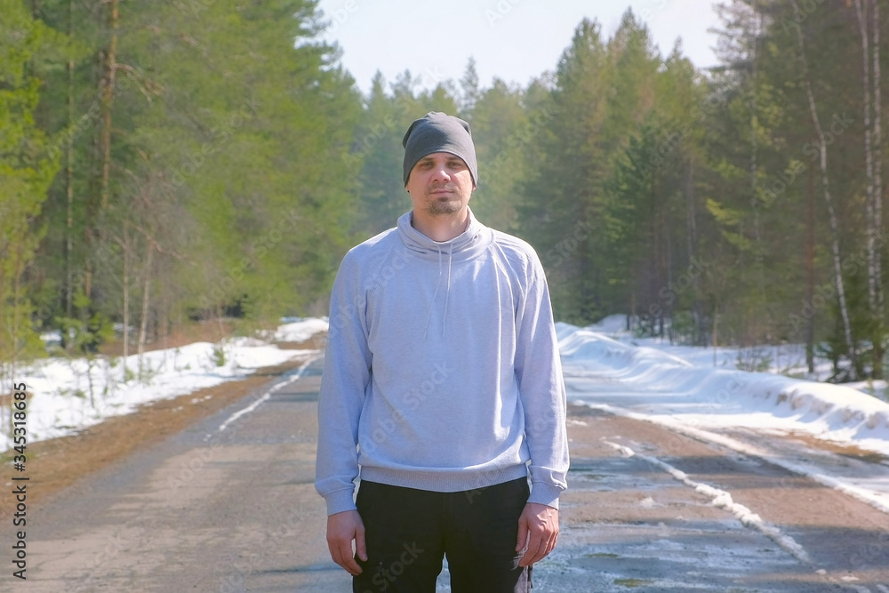 Portrait of man in cap in forest standing on country asphalt road and looking at camera. Warm spring sunny day with melting snow on street. Young caucasian guy is walking on fresh air. Portrait of man in cap in forest standing on country asphalt road and looking at camera. Warm spring sunny day with melting snow on street. Young caucasian guy is walking on fresh air.
