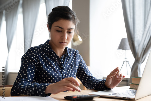 Head shot focused young indian woman calculating domestic expenses, utility bills payments, sitting at table with computer. Concentrated millennial hindu girl managing monthly budget alone at home. photo