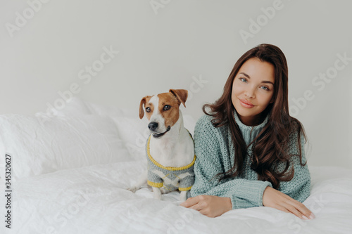 Photo of lovely dark haired woman dresses her dog for cold weather, going to have outdoor walk, lie together at comfortable bed against white background, enjoys relaxation time. Animal care.