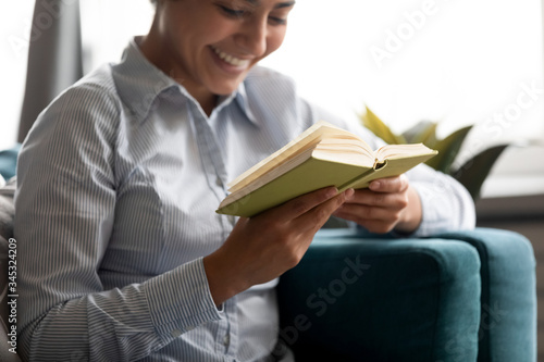 Focus on paper book in hands of happy millennial indian woman. Smiling young hindu ethnic girl reading favorite novel story, resting alone on comfortable couch in living room, hobby pastime concept.
