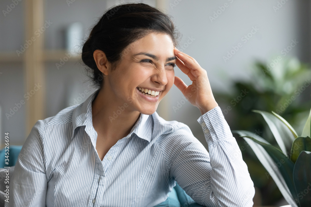 Happy young hindu woman touching forehead, looking away, thinking visualizing future, relaxing on sofa in living room. Head shot millennial indian girl laughing, feeling excited about good news. - obrazy, fototapety, plakaty 