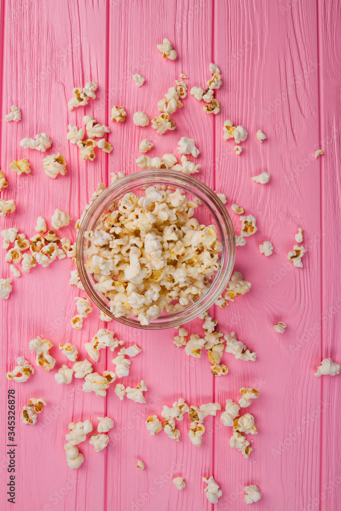 Flat lay popcorn in a glass bowl and sprinkled popcorn around it. Pink wooden desk.