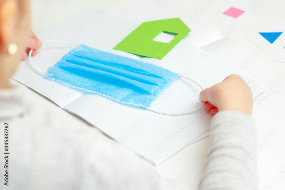 Medical mask in child hands over open school notebook with paper house on the desk. Stay home and education concept. Medical mask in child hands over open school notebook with paper house on the desk. Stay home and education concept.