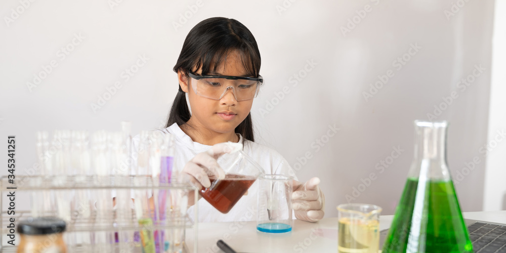 Photo of young adorable school girl holding a chemistry glassware while doing a scientific experiment and sitting at the modern white table with white laboratory wall as background. Photo of young adorable school girl holding a chemistry glassware while doing a scientific experiment and sitting at the modern white table with white laboratory wall as background.