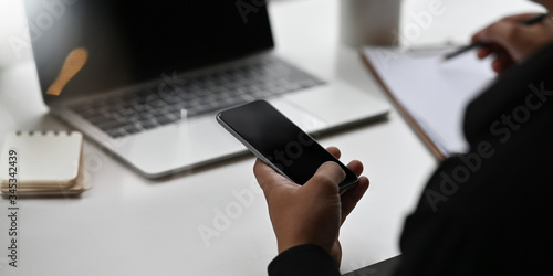 Cropped of businessman's hands holding a smartphone while writing on clipboard and sitting in front computer laptop that putting on white working desk and surrounded by notebook and paper work.