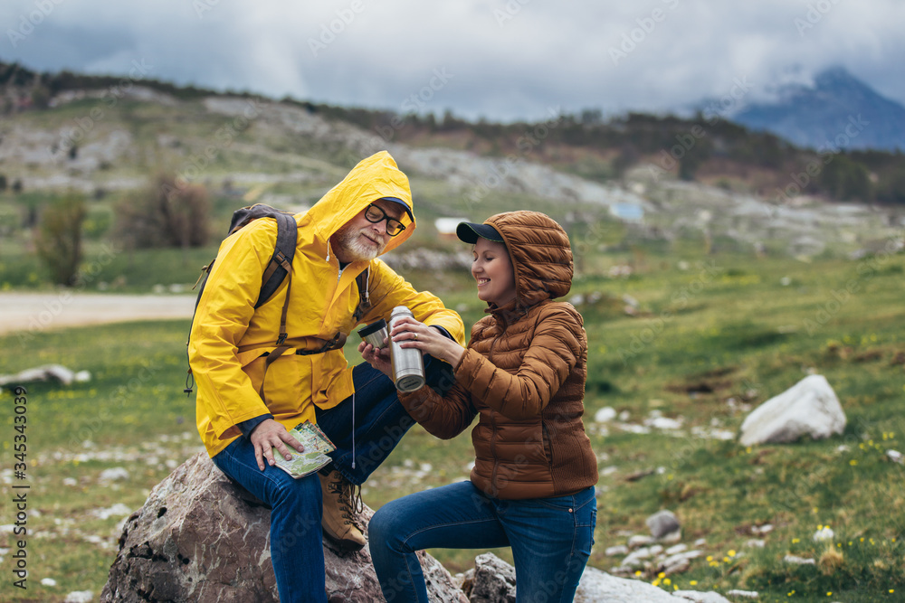 Middle age couple with backpack taking break of hiking in a beautiful nature and taking tea or water from a thermos flask