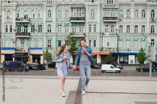 Romantic and happy caucasian couple in casual clothes walking together through the streets. Love, relationships, romance, happiness concept. Man and woman with disposable cups of coffee in the city.