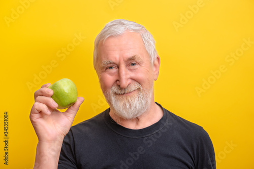 Senior man is giving green apple and 
 showed big finger over yellow background. Senior man take morning