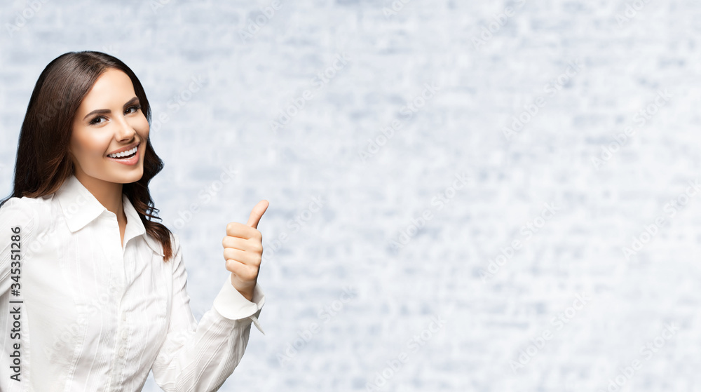 Portrait of happy smiling young excited businesswoman, showing thumb up hand sign gesture. Success in business concept studio shot. Standing against white brick wall background. Portrait of happy smiling young excited businesswoman, showing thumb up hand sign gesture. Success in business concept studio shot. Standing against white brick wall background.
