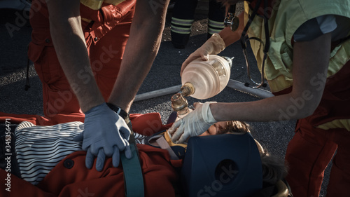 On the Car Crash Traffic Accident Scene: Paramedics Saving Life of a Female Victim who is Lying on Stretchers. They Apply Oxygen Mask, Do Cardiopulmonary Resuscitation / CPR and Perform First Aid