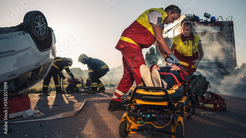 On the Car Crash Traffic Accident Scene: Paramedics Saving Life of a Female Victim who is Lying on Stretchers. They Apply Oxygen Mask, Do Cardiopulmonary Resuscitation / CPR and Perform First Aid photo
