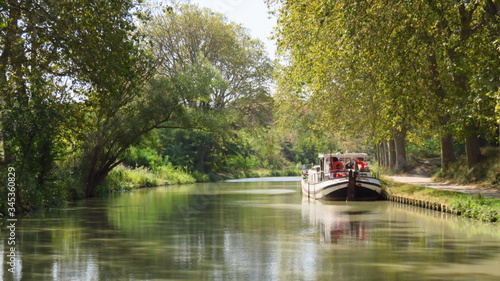 Stampa su tela Tourisme fluvial, péniche amarrée sur le Canal du Midi bordé d'arbres en été (Fr