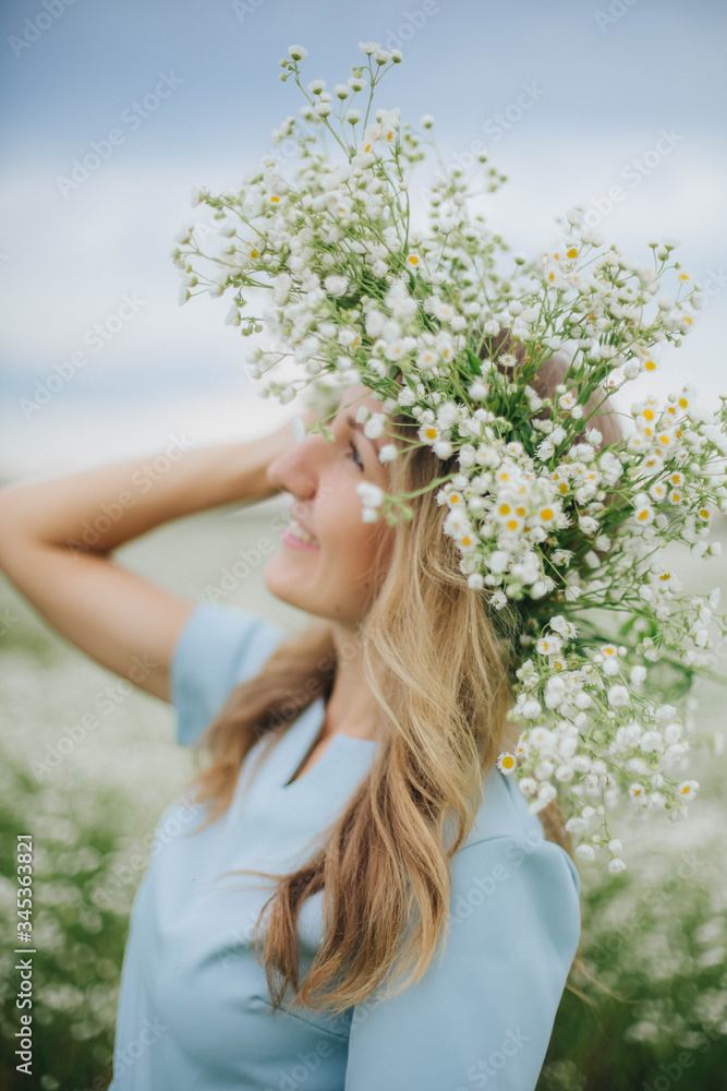 Fototapeta premium beautiful blonde girl in a field of daisies. wreath of wildflowers on his head. woman in a blue dress in a field of white flowers. charming girl with a bouquet of daisies. summer tender photo
