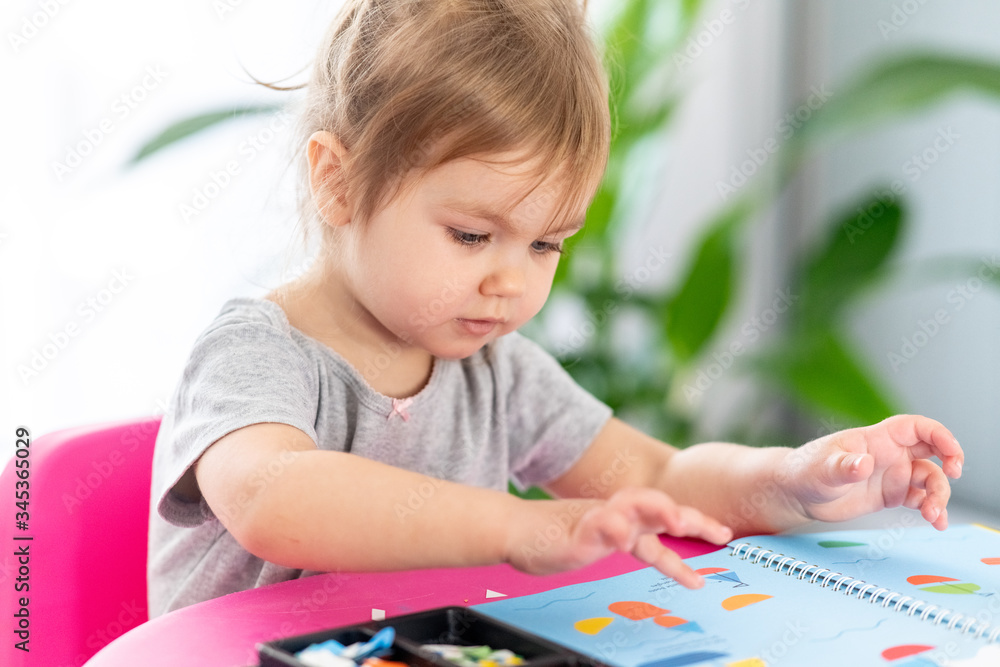 Concentrated cute 2year old girl is playing with stickers at home. Quarantine. Stay home. Indoor activities. Concentrated cute 2year old girl is playing with stickers at home. Quarantine. Stay home. Indoor activities.