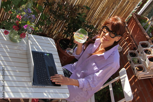 Donna lavora da casa sul terrazzo con computer, drink e fiori in una bella giornata di sole photo