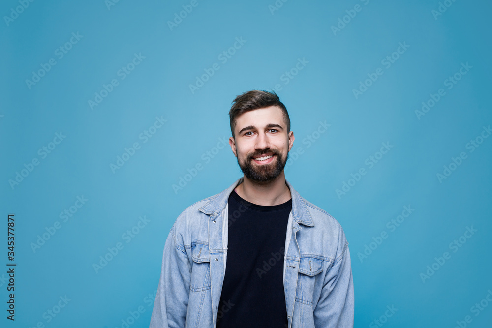 Young cheerful man in black t-shirt and denim jacket. Guy with beard expresses happiness. Blue background. Young cheerful man in black t-shirt and denim jacket. Guy with beard expresses happiness. Blue background.